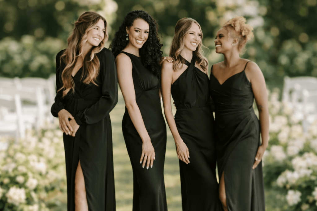 Four women in black dresses standing outdoors with greenery and flowers in the background