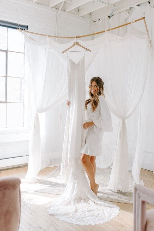 Bride stands in Kennedy Blue robe with wedding dress hanging next to her